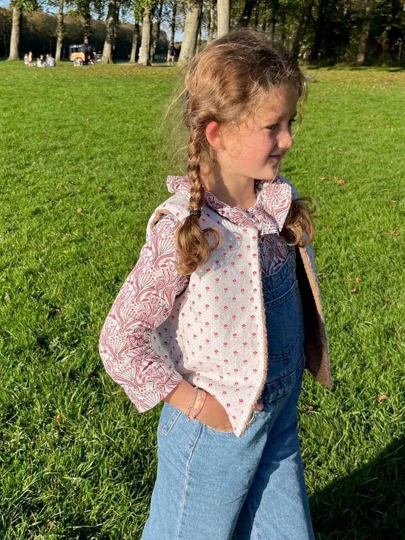 Fillette de profil dans un parc, gilet en jean et blouse fleurie rose à col volanté — vêtements enfant fait main By Mésange