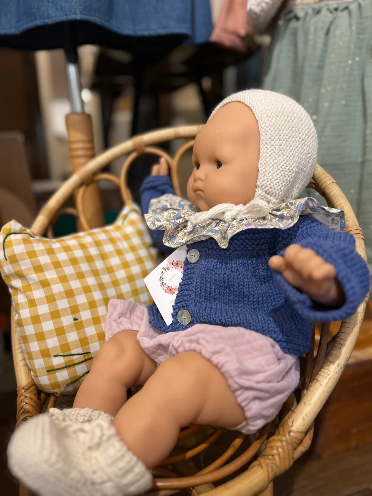 Poupée habillée By Mésange Poupée habillée By Mésange — cardigan bleu, col fleuri et bloomer rose dans un fauteuil en rotin